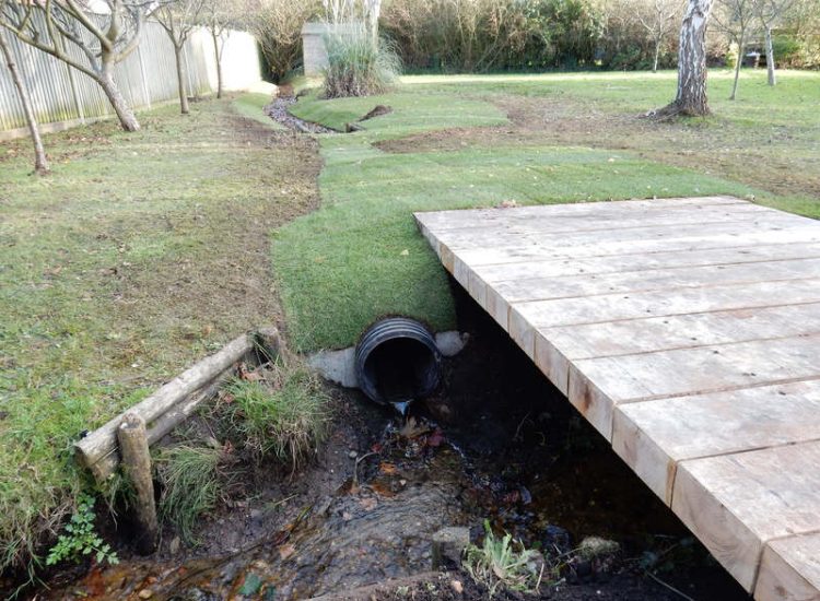 Garden Drainage and Brook Management Worplesdon, Guildford, Surrey - An existing brook had become over grown and blocked with vegetation and silt build up. Brook was cleared, re-ditched, a new under ground pipe installed, surrounding banks and lawns re-turfed and finally construction of a new Oak sleeper bridge to replace the old one. Garden Drainage - MDB Landscapes